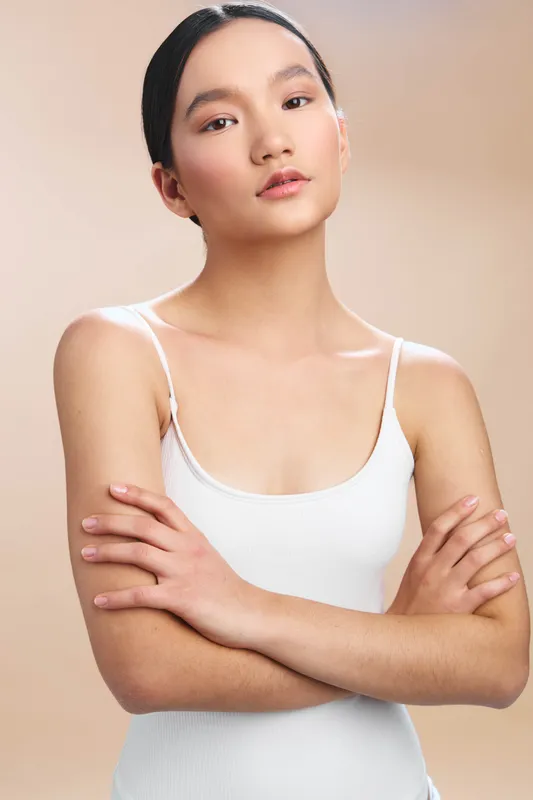 Asian woman poses in a studio portrait wearing a white top against a soft beige backdrop. She appears confident and calm, exuding poise, elegance, and focus in a clean, minimalist setting.