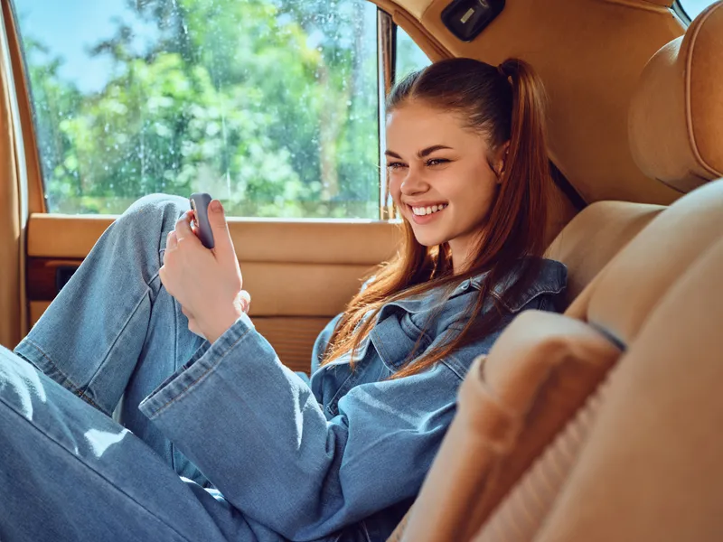 Young woman in denim outfit smiling while using smartphone inside a car, enjoying a sunny day, showcasing happiness and casual lifestyle.