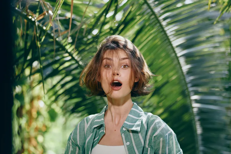 Woman in a casual striped shirt stands among lush tropical foliage, astonished expression, wide eyes, open mouth, surprised mood.