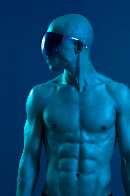 Athletic man with blue body paint wearing futuristic sunglasses, captured in a vibrant light. The image reflects an edgy aesthetic and contemporary artistic trend.