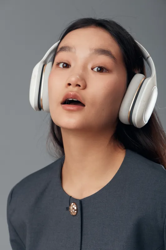 Young woman wearing large wireless headphones looking slightly surprised or curious with parted lips and dark long hair against neutral background for music or technology concept.