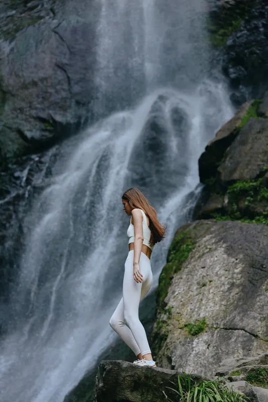 woman standing near waterfall in white athletic wear, natural outdoor setting, long brown hair, serene mood, rocky landscape, lush greenery