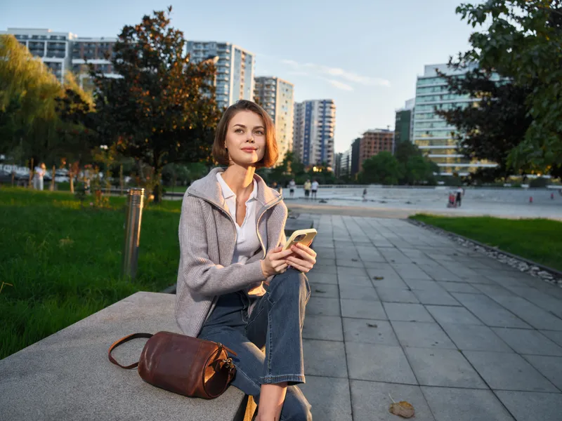 A cheerful professional sits outdoors in an urban park, texting on a smartphone while smiling, creating a friendly, energetic vibe in a modern cityscape.