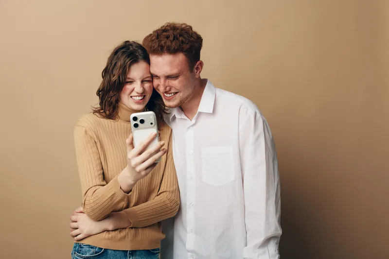 Happy young couple with curly hair takes selfie together on smartphone. They smile and embrace in casual clothes against beige background. Concept of friendship, technology, and joy.