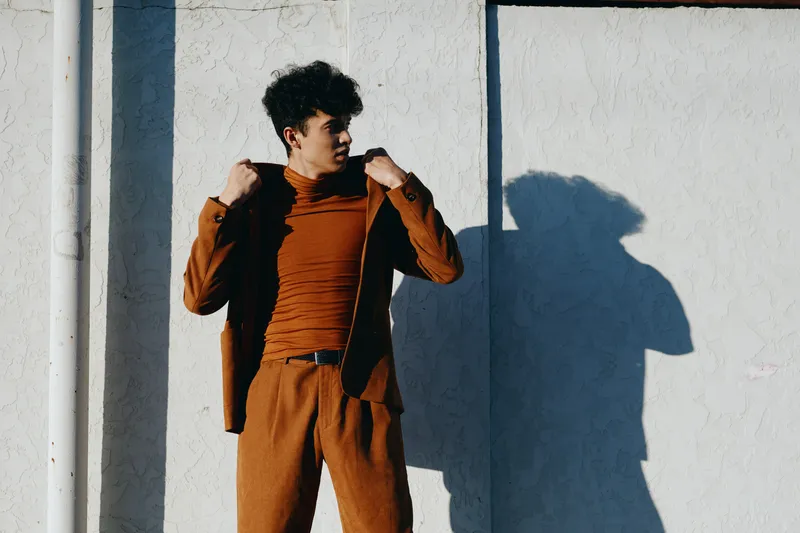 Модный молодой человек в коричневом наряде стоит на фоне белой стены, что подчёркивает его уверенную осанку и современный стиль
