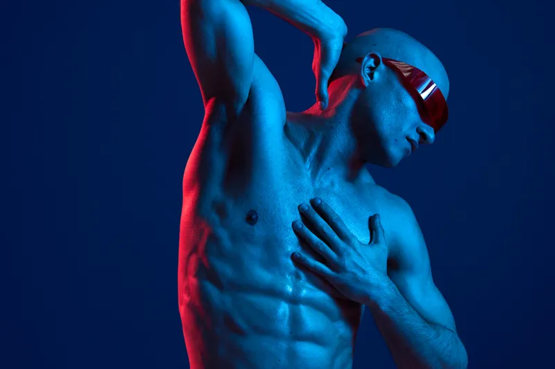 Muscular man posing dramatically against a blue and red backdrop, showcasing strength and confidence. This is a striking color image capturing dynamic movement.