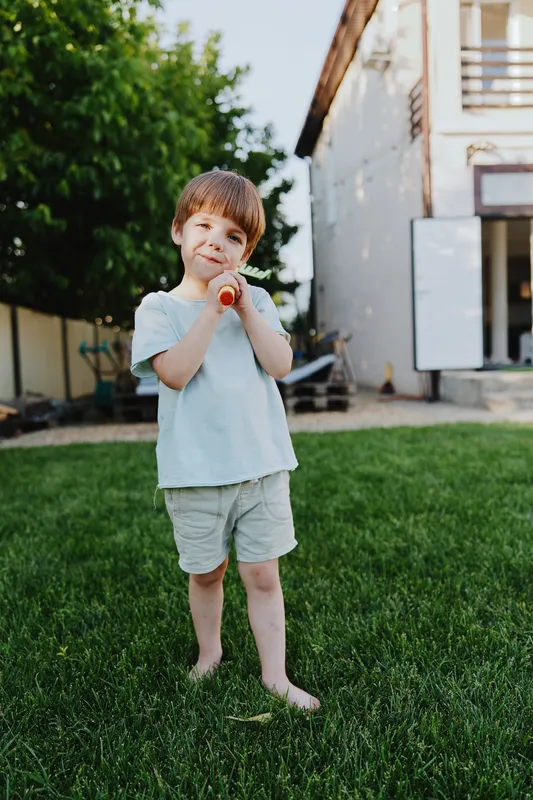 Happy boy holding a carrot, enjoying a sunny day outdoors in a lush green backyard with a playful atmosphere and a carefree spirit.