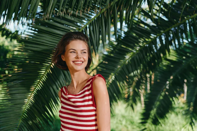 A cheerful woman in a red striped top stands amid lush palm leaves outdoors, smiling warmly, radiating relaxed confidence and sunny energy, natural charm.