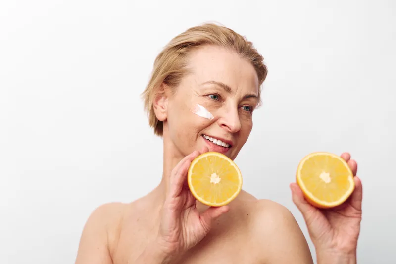 Mature woman with short hair holds two orange halves near her face, smiling with natural skin and white cream on cheek. Concept of healthy lifestyle, skincare, citrus fruit, and wellness.