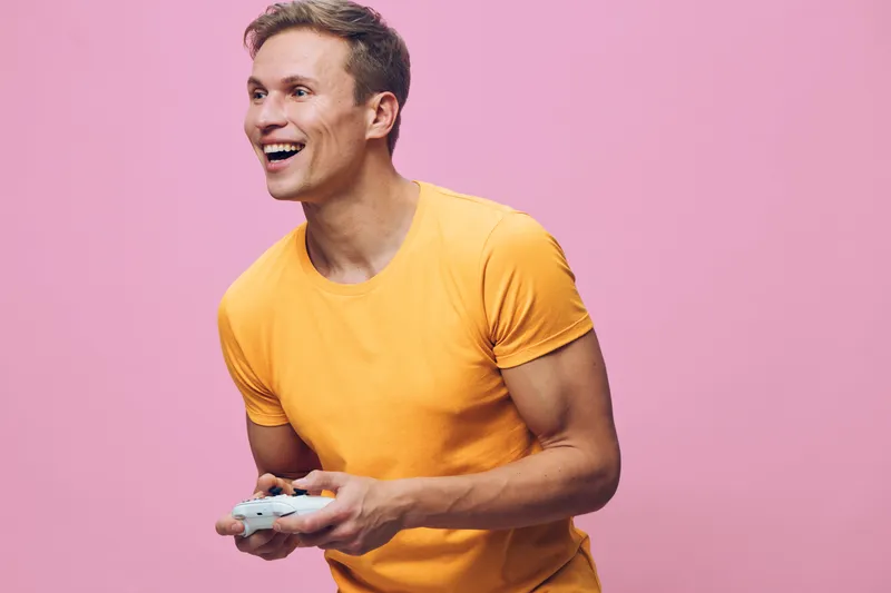Smiling man in casual yellow t-shirt playing video games holding game controller isolated on pink background. Male leisure activity, happiness, entertainment, relaxed lifestyle concept.