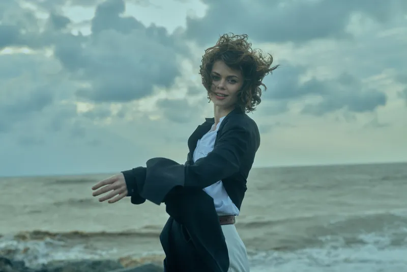 Woman on a windy beach adjusting a coat, ocean breeze and cloudy sky, casual chic mood, outdoor fashion scene, motion and freedom by the shore.