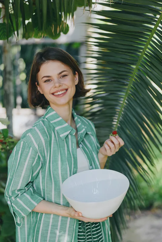Smiling woman outdoors holding a white ceramic bowl, wearing a green striped shirt, with lush palm leaves in the background.