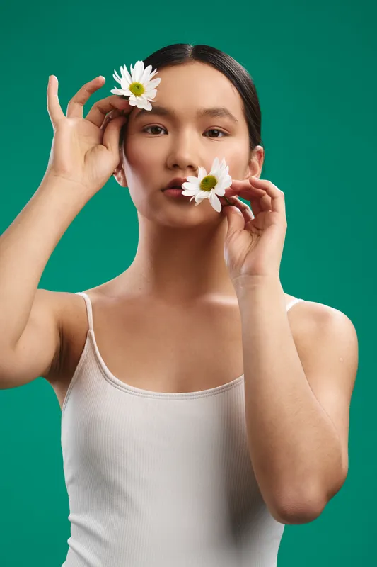 Asian woman poses with flowers in a fresh studio portrait, wearing a light tank top against a solid green background, creating a gentle, natural and stylish mood.