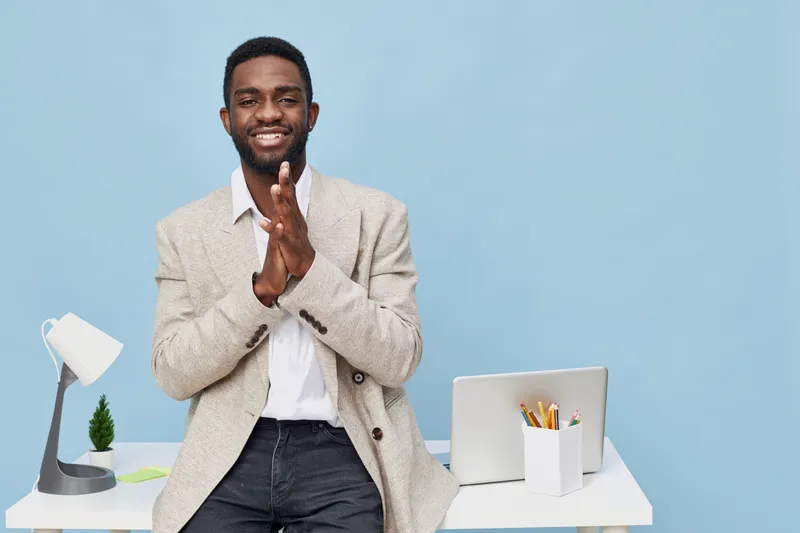 Happy smiling man sitting at modern desk in bright office environment, holding hand in greeting gesture, with laptop, stationery, and lamp on background, casual business attire, professional lifestyle