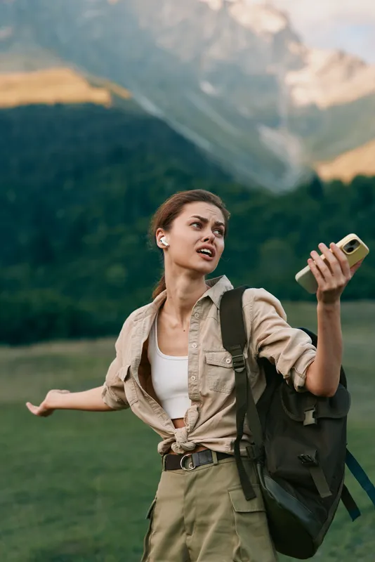 A young woman in casual outdoor clothing holds a smartphone and gestures with her hand while standing in a green mountainous landscape. Lifestyle concept outdoors.