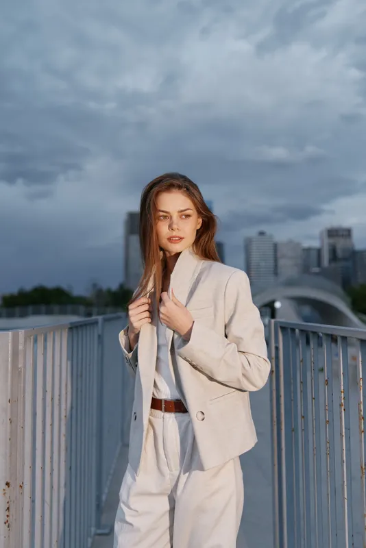 Confident woman in light business attire poses on a bridge against a skyline backdrop, exuding professionalism and modernity in an urban setting.