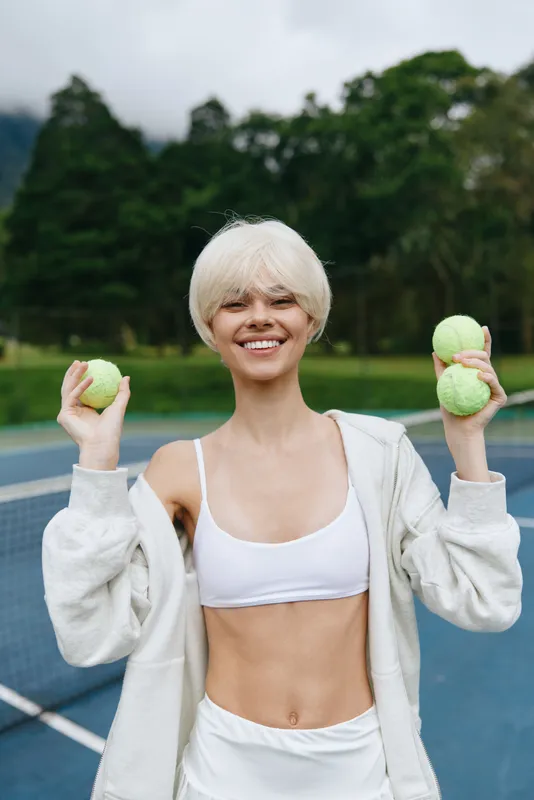Young woman with short blonde hair smiling joyfully while holding tennis balls on a sunny day, dressed in sporty attire, showcasing a healthy lifestyle