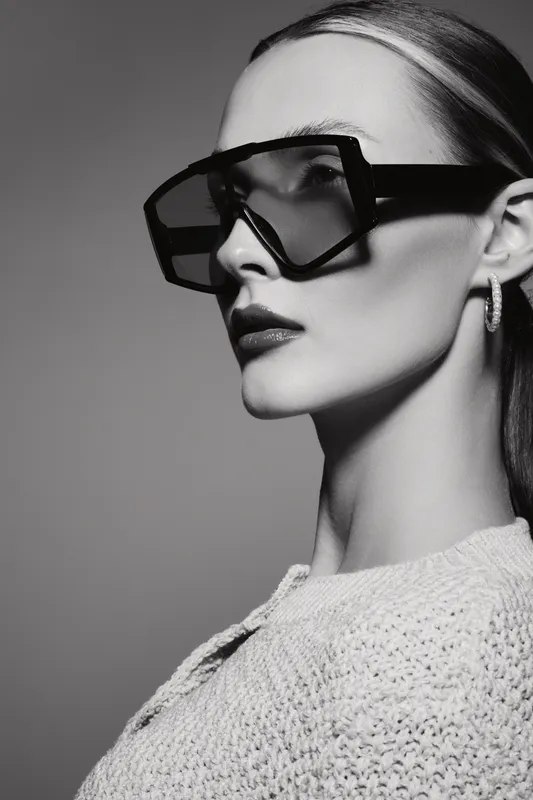 Portrait of a young white woman with pale skin wearing oversized glasses and earrings in a knitted sweater. Studio fashion concept with monochrome lighting.