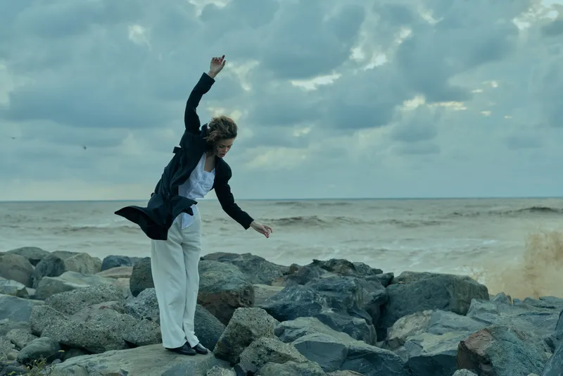 Woman on a rocky shoreline performing a dynamic leap, jacket flaring, sea and dramatic sky behind, capturing motion, style and energy in an outdoor coastal scene.