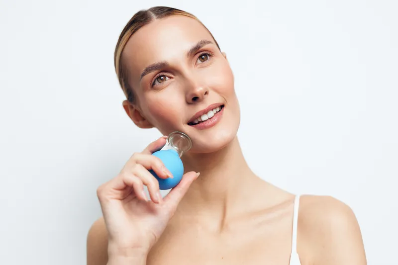 Young woman with clear skin holding blue cosmetic bottle near face, smiling softly, natural makeup, bright background, skincare routine, beauty concept, fresh and healthy look
