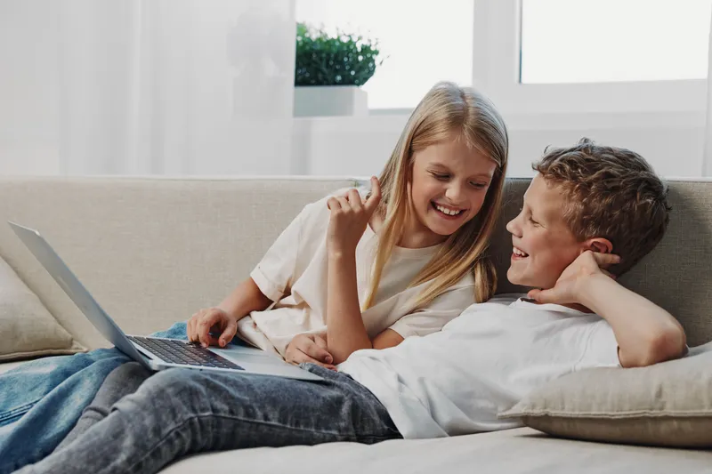 Happy children lying on sofa with laptop smiling and interacting together in bright living room. Concept of friendship, technology, leisure, and childhood bonding at home with casual clothing.