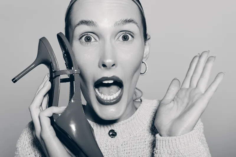 Surprised Caucasian woman with light skin holds high heel shoe near face, open mouth and wide eyes expressing shock or excitement in studio.