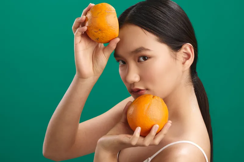 an asian woman holds oranges near her face against a solid green background, smiling softly and creating a fresh, vibrant portrait with a playful mood.