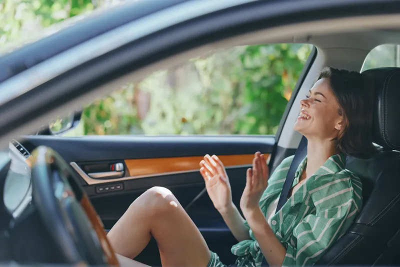 A joyful woman sits in a car seat, smiling and laughing, enjoying a relaxed moment inside a modern vehicle.