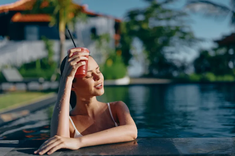 A woman relaxes at a sunny poolside, holding a red drink up to the light. Clear water, warm glow, and casual summer mood convey leisure, self care, and carefree escape.
