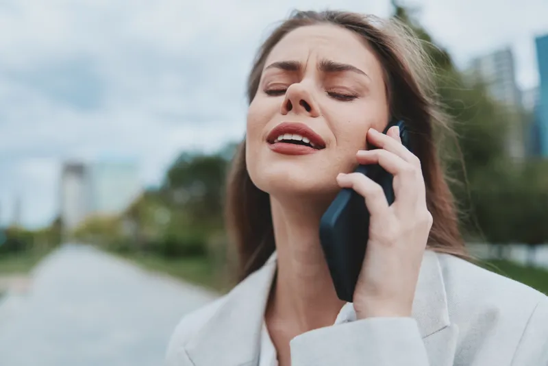 Young woman in business attire experiencing a moment of stress while talking on the phone in a vibrant urban park during the day.
