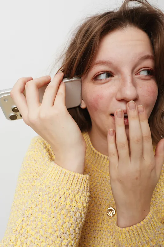 Young woman in yellow sweater holds smartphone to ear and covers mouth with hand, smiling shyly. Concept of communication, surprise, casual lifestyle, and positive emotions in everyday life.
