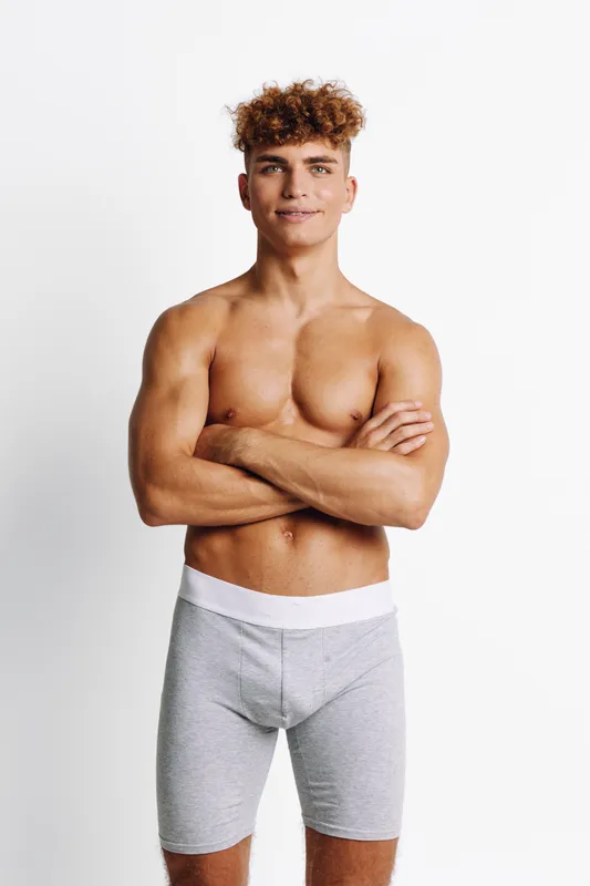 Young man with curly hair standing shirtless with arms crossed wearing gray boxer briefs against white background. Confident male model posing with smile and athletic body.