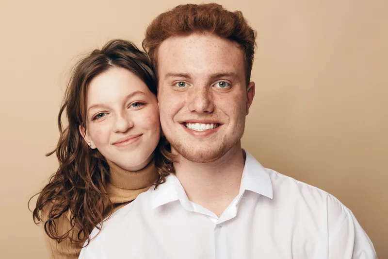 Smiling young couple with red hair and curly brown hair posing together against beige background. Happy man and woman show affection and warmth in casual clothing. Concept of love and friendship.