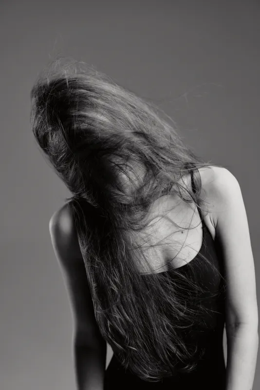 Black and white portrait of a young woman with flowing hair in motion, wearing a dark tank top, expressing mystery and movement.