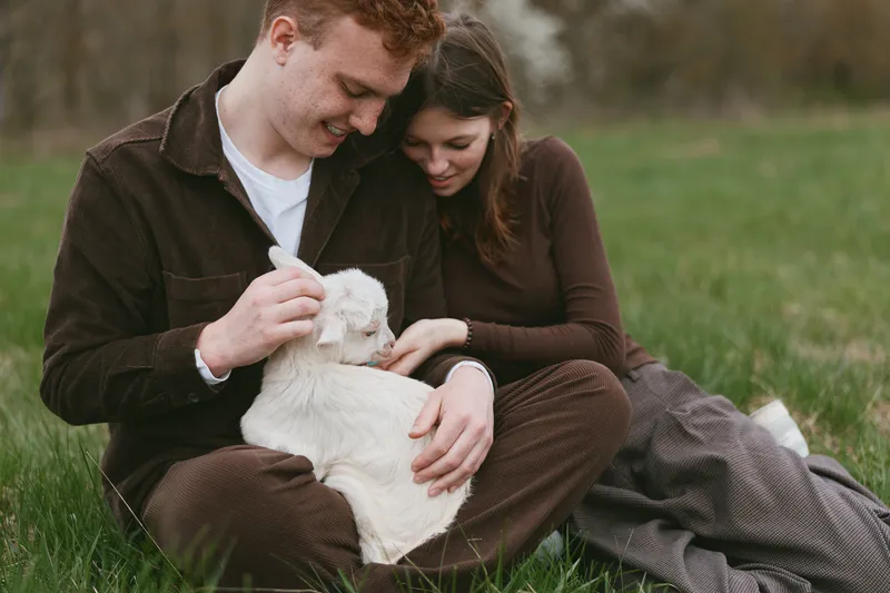 Loving couple outdoors with a white goat kid, sitting on grass, gentle interaction, candid lifestyle moment, warmth and connection, natural light, tender companionship, rural calm
