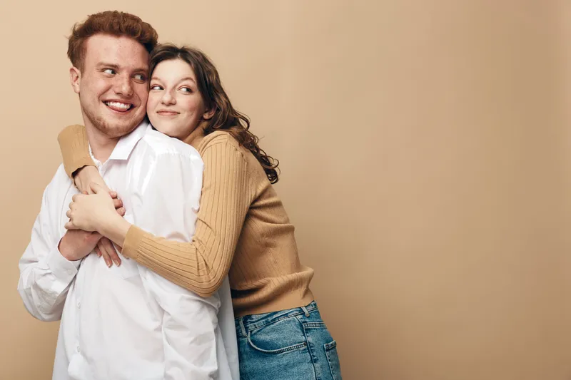 Happy young couple embraces and smiles together on beige background. Casual clothing and warm mood create concept of love, friendship, and relationship in studio setting.