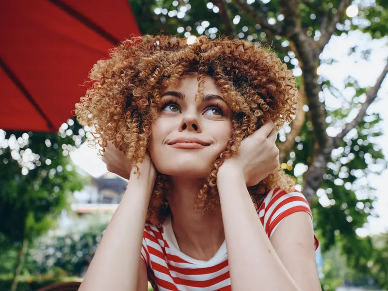 Portrait of a joyful young woman with curly hair resting her chin on her hands, outdoors under a red umbrella with greenery, capturing a dreamy, hopeful mood.