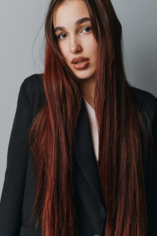 Woman with long straight brown hair and natural makeup wearing black blazer posing against grey background in studio. Portrait of young female model with serious expression and casual style.