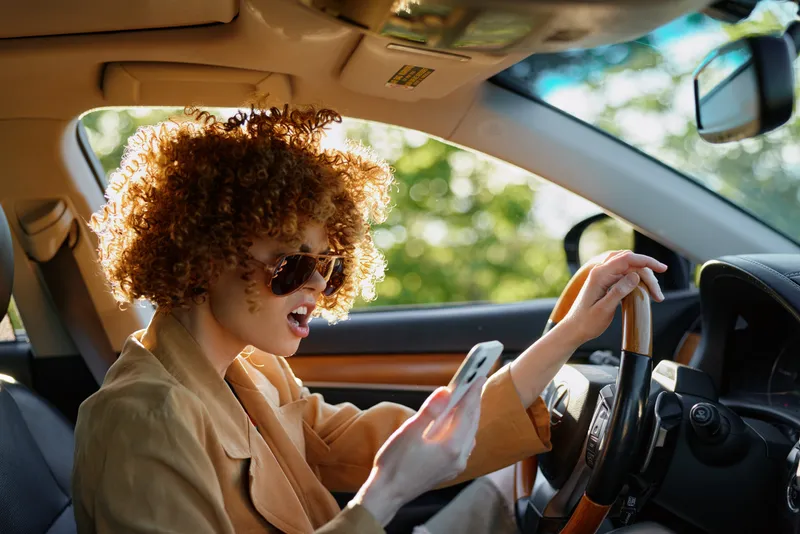Cheerful woman driving a car, glancing at her phone, wearing sunglasses, curly hair in sunlight, capturing a casual lifestyle moment inside a warm, vibrant setting