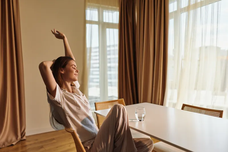 Woman relaxing at home in the morning, stretching back in a chair by a dining table with sunlight through curtains, casual loungewear and a glass of water nearby.