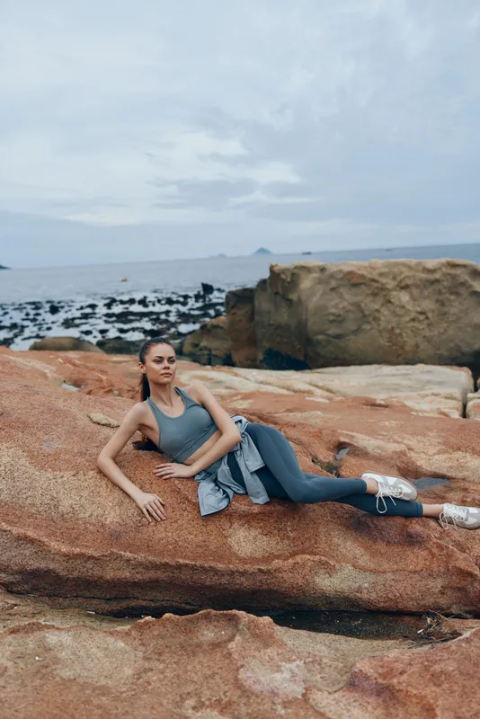Young woman in athletic wear relaxing on rocky beach, embodying fitness and serenity amidst natural landscape, perfect for healthy lifestyle concepts