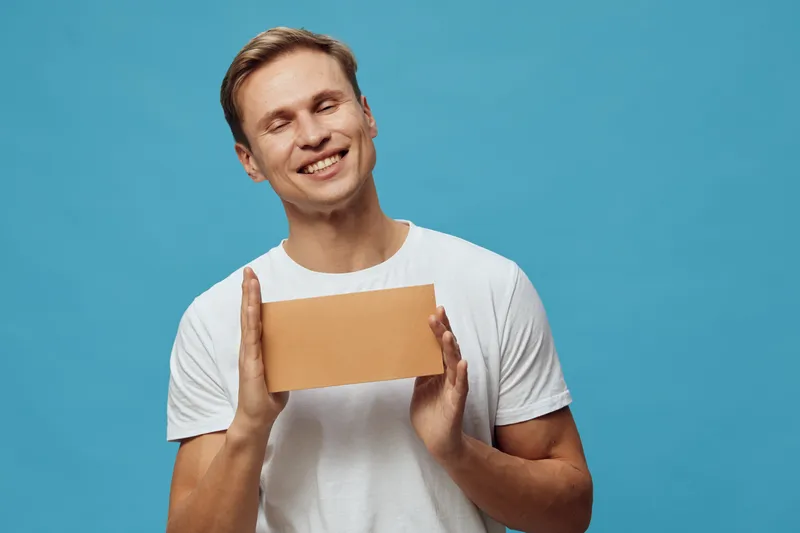 Smiling happy young man holding cardboard box with closed eyes isolated on blue background studio portrait. People lifestyle concept, delivery, parcel, package, gift, excitement, happiness