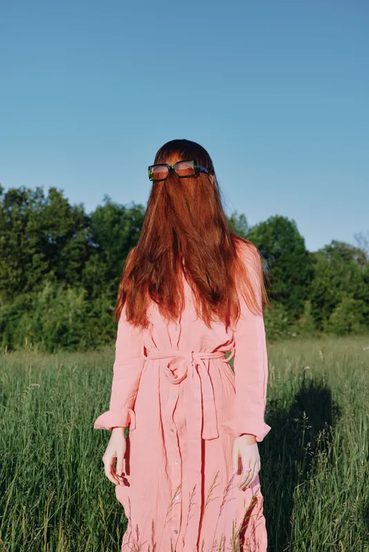 A woman with long red hair obscuring her face stands in a sunny green field wearing a pink dress, conveying a sense of mystery and serenity