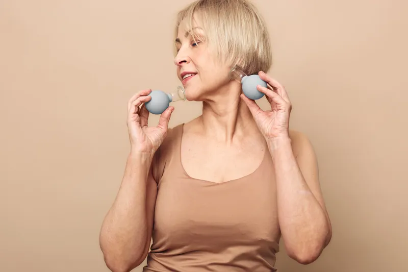 Mature woman with short blonde hair uses facial massage tools on neck and cheeks in beige tank top. Concept of anti aging skincare, self care, wellness, and natural beauty at home.