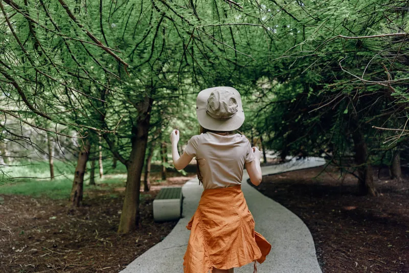 Woman walking on a curved park path under tall trees, wearing a sun hat and orange skirt, back view of a traveler enjoying nature and a peaceful woodland stroll.