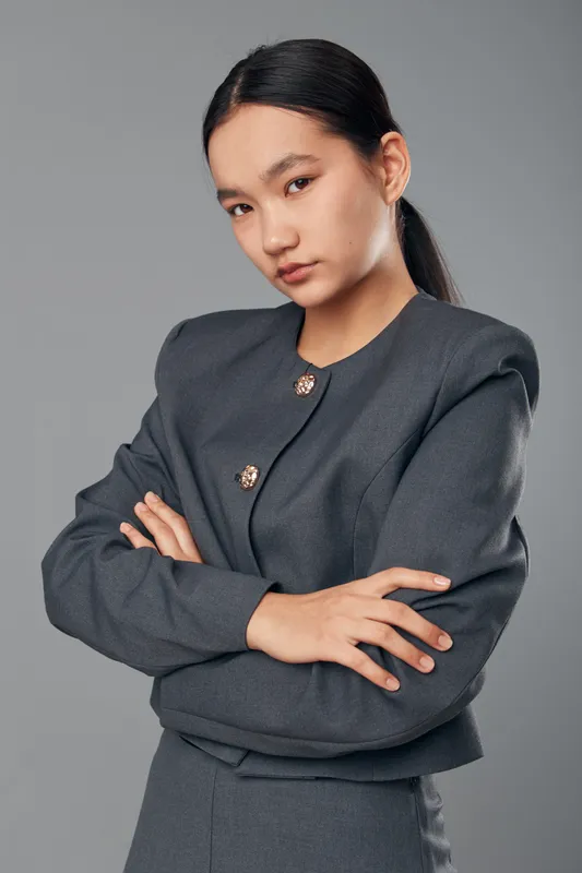 Young woman with long dark hair and crossed arms wearing a gray formal jacket posing confidently on gray background. Stylish professional with serious expression and natural makeup.