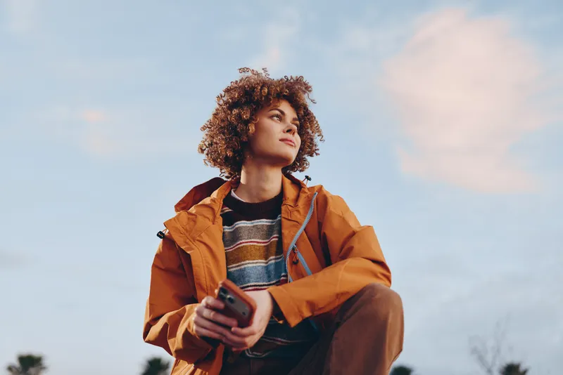 Curly haired woman in orange jacket holding a smartphone outdoors, looking away, lifestyle moment in warm sunset light, travel style, casual communication, soft sky background, confident mood