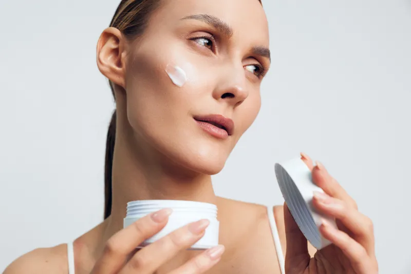 Woman skincare close up, applying cream to face with cotton, checking mirror for smooth skin, beauty routine with hygiene focus, gentle cosmetics treatment, calm studio light, self care