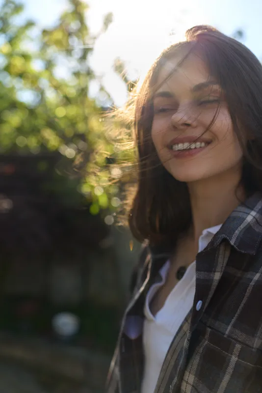 woman outdoors lifestyle portrait, smiling, casual shirt in sunlight, relaxed pose, gentle breeze, natural greenery backdrop, warm colors, candid moment