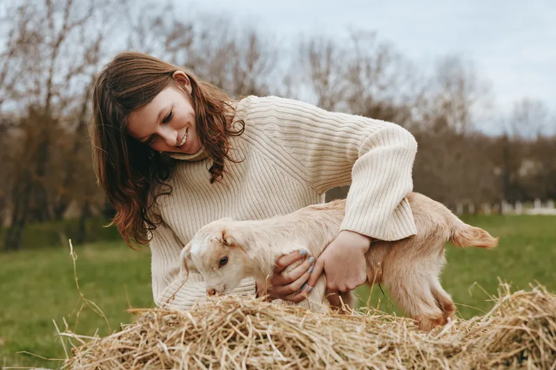 Joyful woman outdoors, gently holding a goat kid on hay, warm natural light, cozy sweater, rustic farm setting, tender moment of connection and care, nature vibes